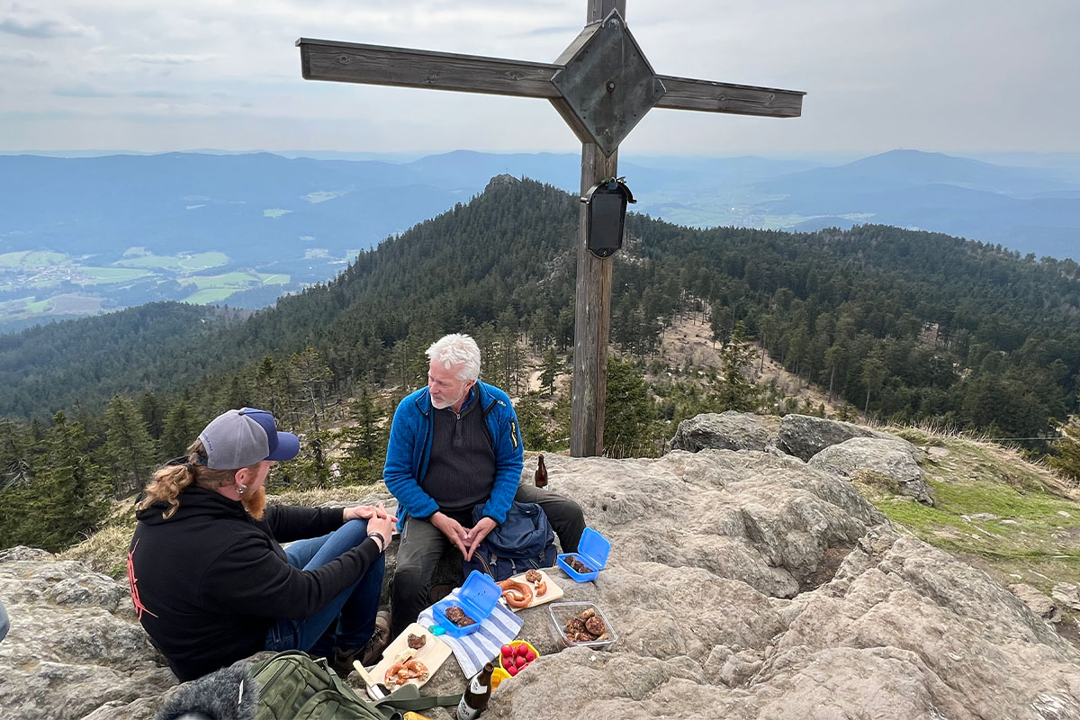 Luki Maurer und Werner Schmidtbauer sitzen unterm Gipfelkreuz bei einer Brotzeit zusammen