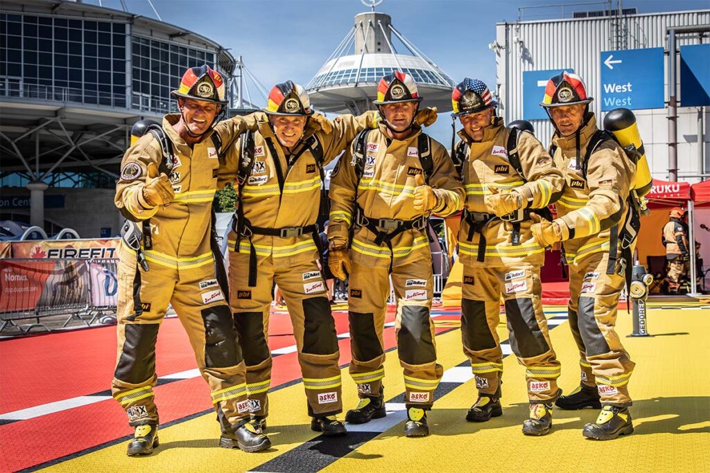 Die Wild 50's stehen in voller Feuerwehrmontur auf dem rot-gelben FireFit Parcours