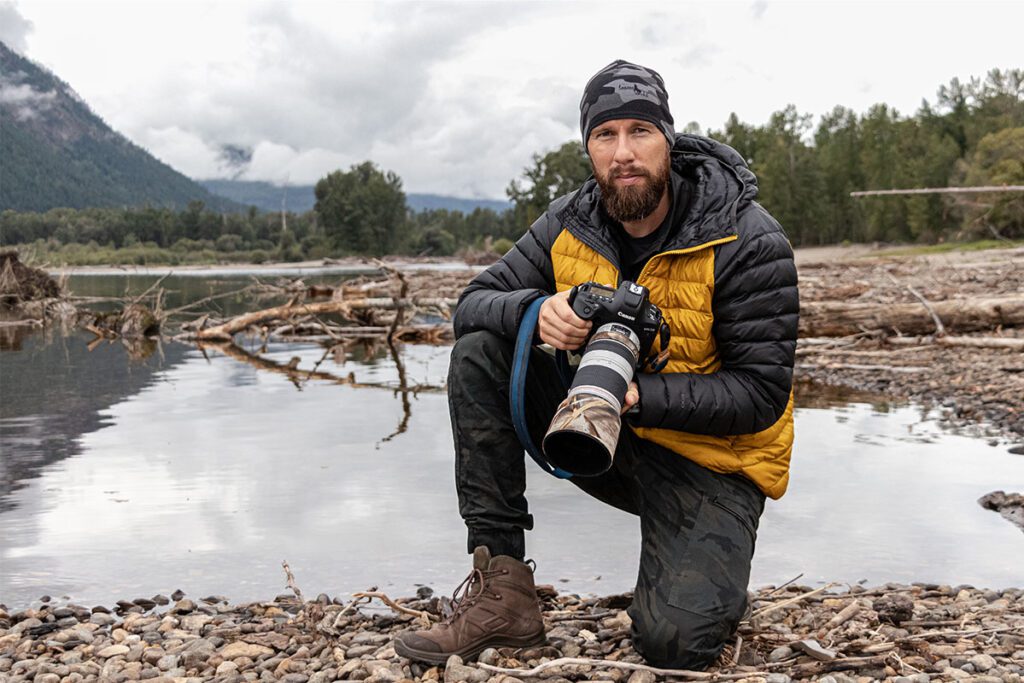 Robert Marc Lehmann kniet mit Kamera in der Hand an einem Flussufer